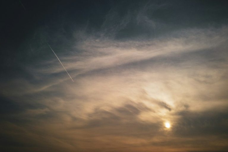 Cielo con nubes dispersas y un sol tenue, acompañado de una estela de avión.
